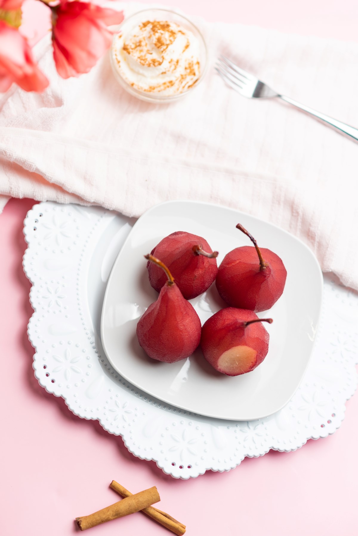 Red wine poached pears on a white plate with cinnamon sticks and a bowl of whipped cream beside the plate.