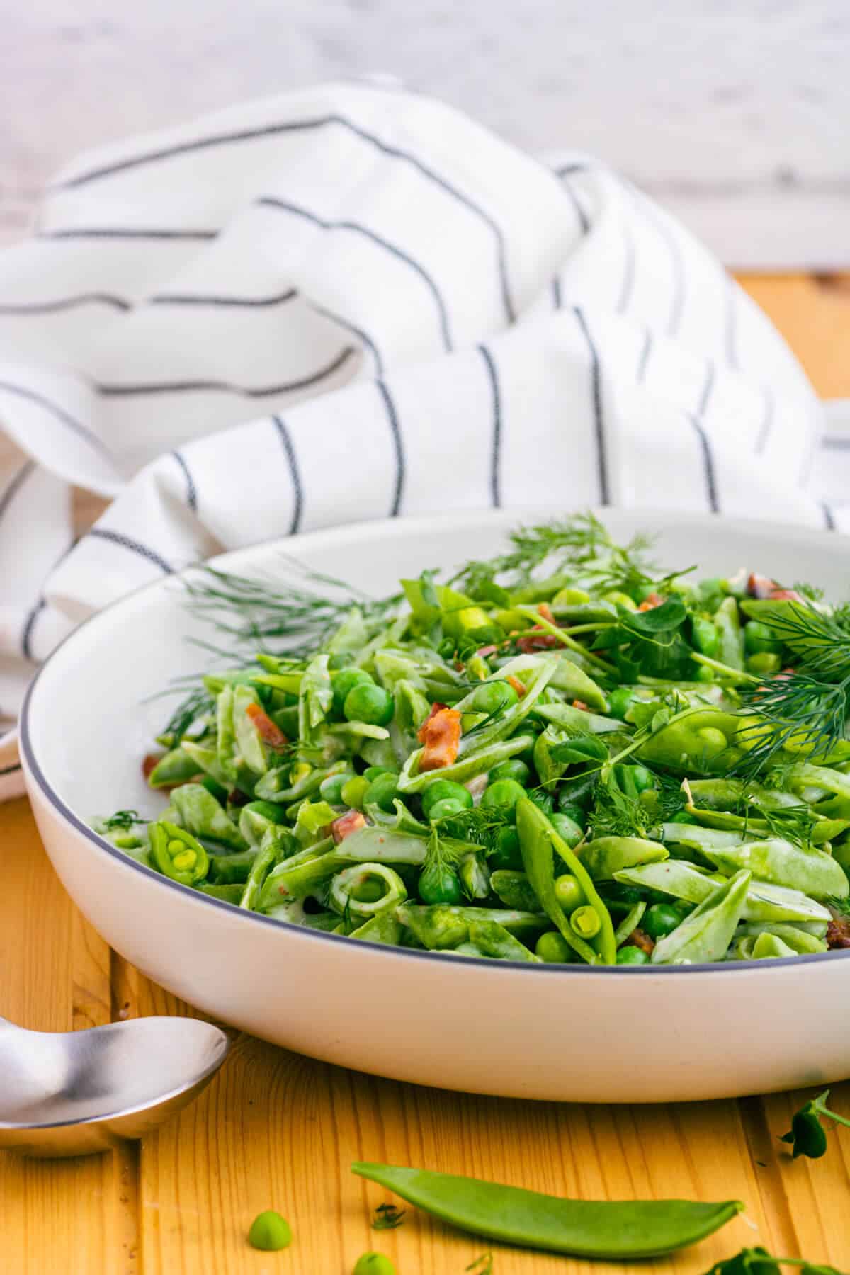 A white bowl filled with bright green sugar snap peas, Spring peas, and bacon with fresh green herbs in a cream sauce.