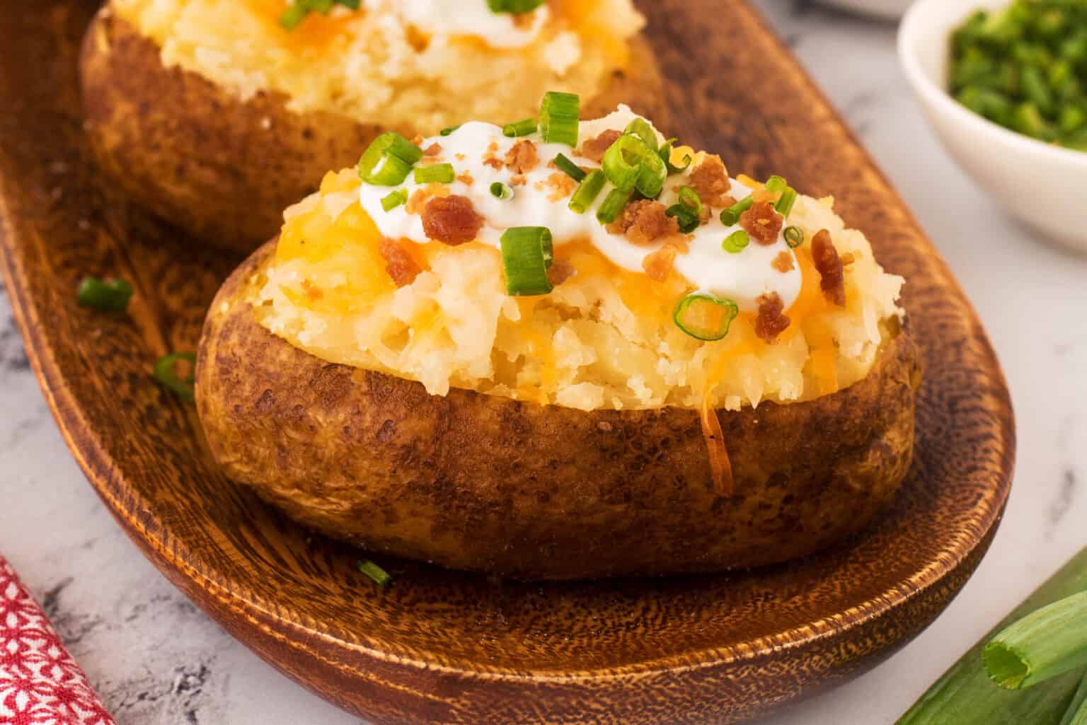A close-up of a stuffed baked potato on a wooden serving plate. The potato is topped with melted cheese, sour cream, chopped green onions, and bits of crispy bacon. In the background, there is a small bowl of chopped green onions and a red patterned cloth.
