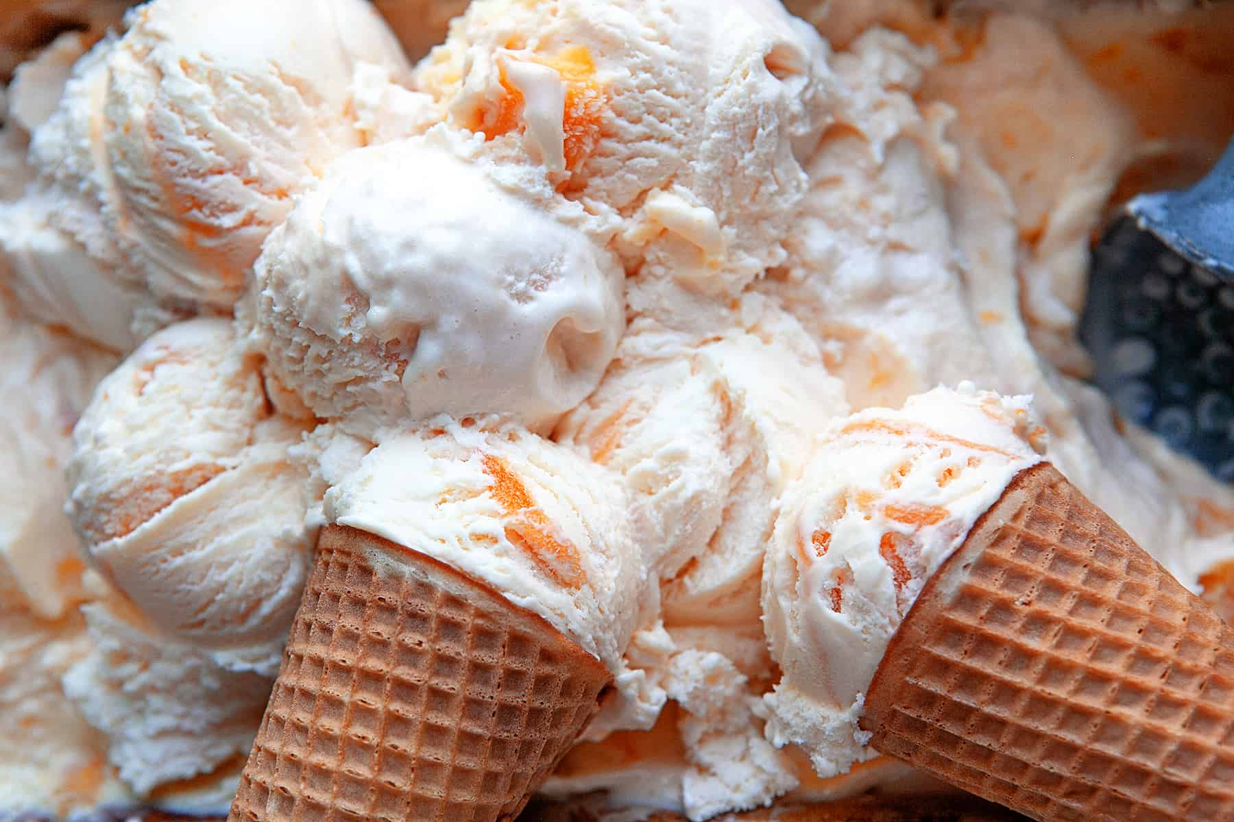 Horizontal photo of balls of peach ice cream and cones in a container. 