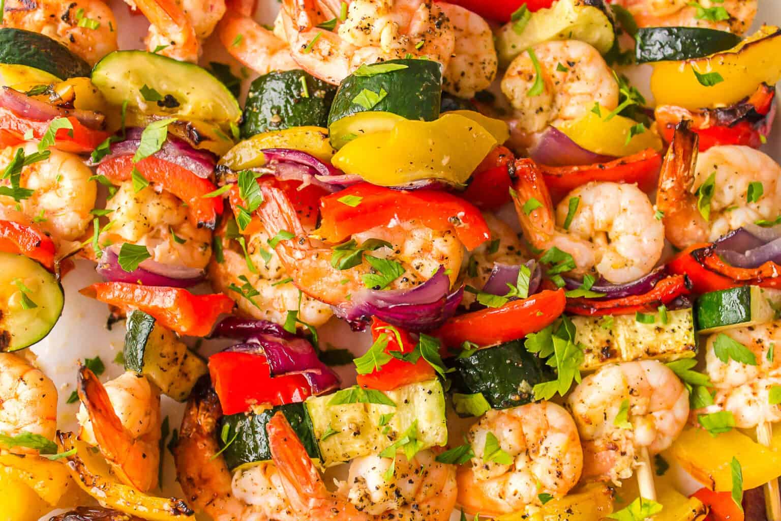 A vibrant display of grilled shrimp and vegetable skewers featuring colorful bell peppers, zucchini, red onions, and garnished with fresh herbs. The shrimp and vegetables are seasoned with spices, appearing fresh and appetizing.