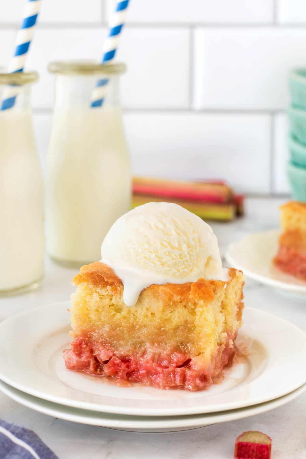 Old-Fashioned Rhubarb Pudding Cake on a plate with ice cream on top. Milk bottles with straws in the background. 