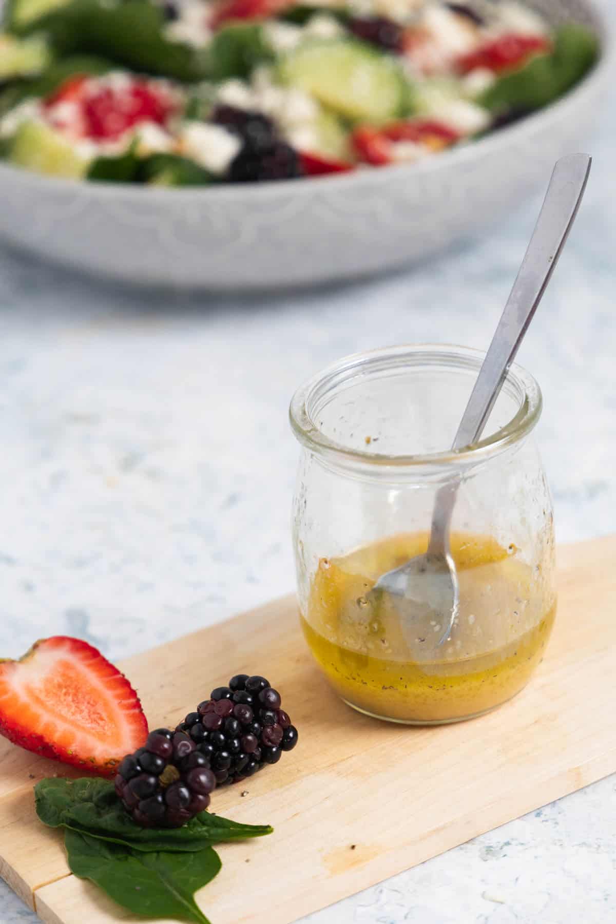glass jar with dressing and a spoon on a wooden board with half a strawberry and a blackberry. 