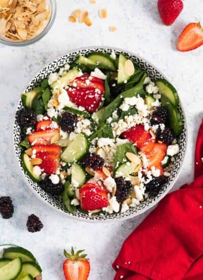 Spinach and strawberry salad in a round dish with some sliced strawberries on the side, toasted almonds and a red napkin.