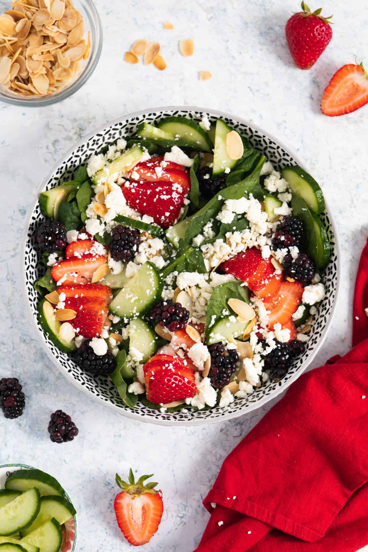 Spinach and strawberry salad in a round dish with some sliced strawberries on the side, toasted almonds and a red napkin.
