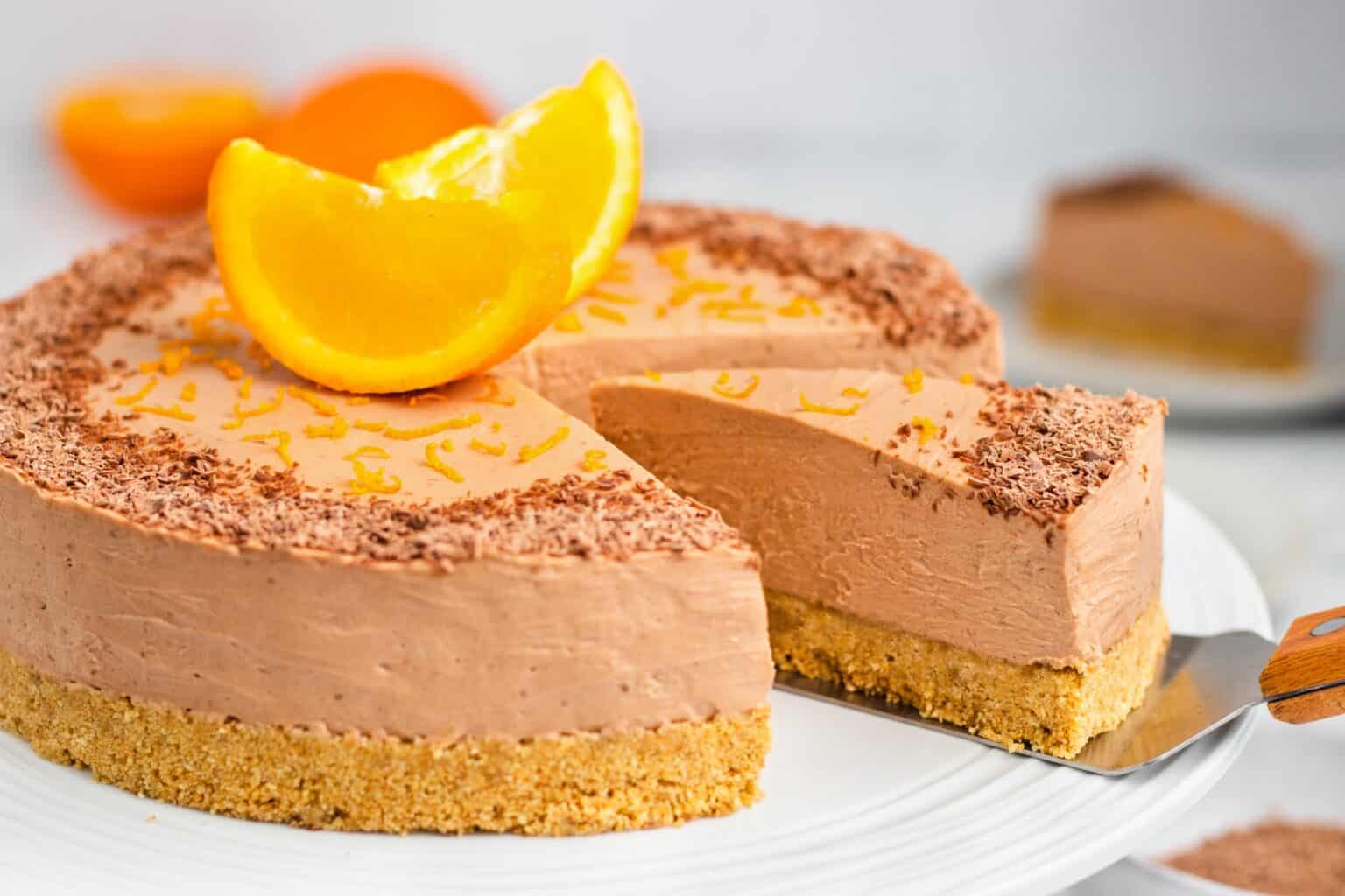 A creamy chocolate cheesecake topped with orange zest and a slice of orange, sitting on a graham cracker crust. A piece is being cut and lifted on a spatula.