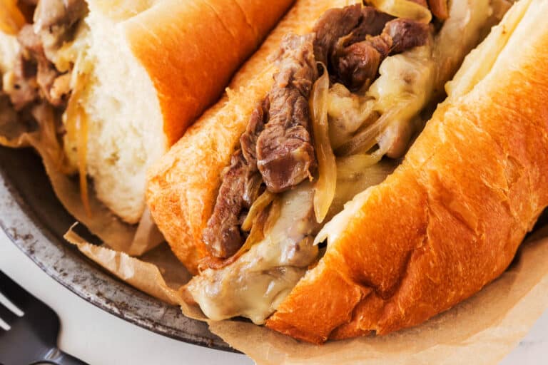 Two warm, crusty hoagie rolls filled with sliced steak, melted cheese, and sautéed onions, served on parchment in a metal tray.