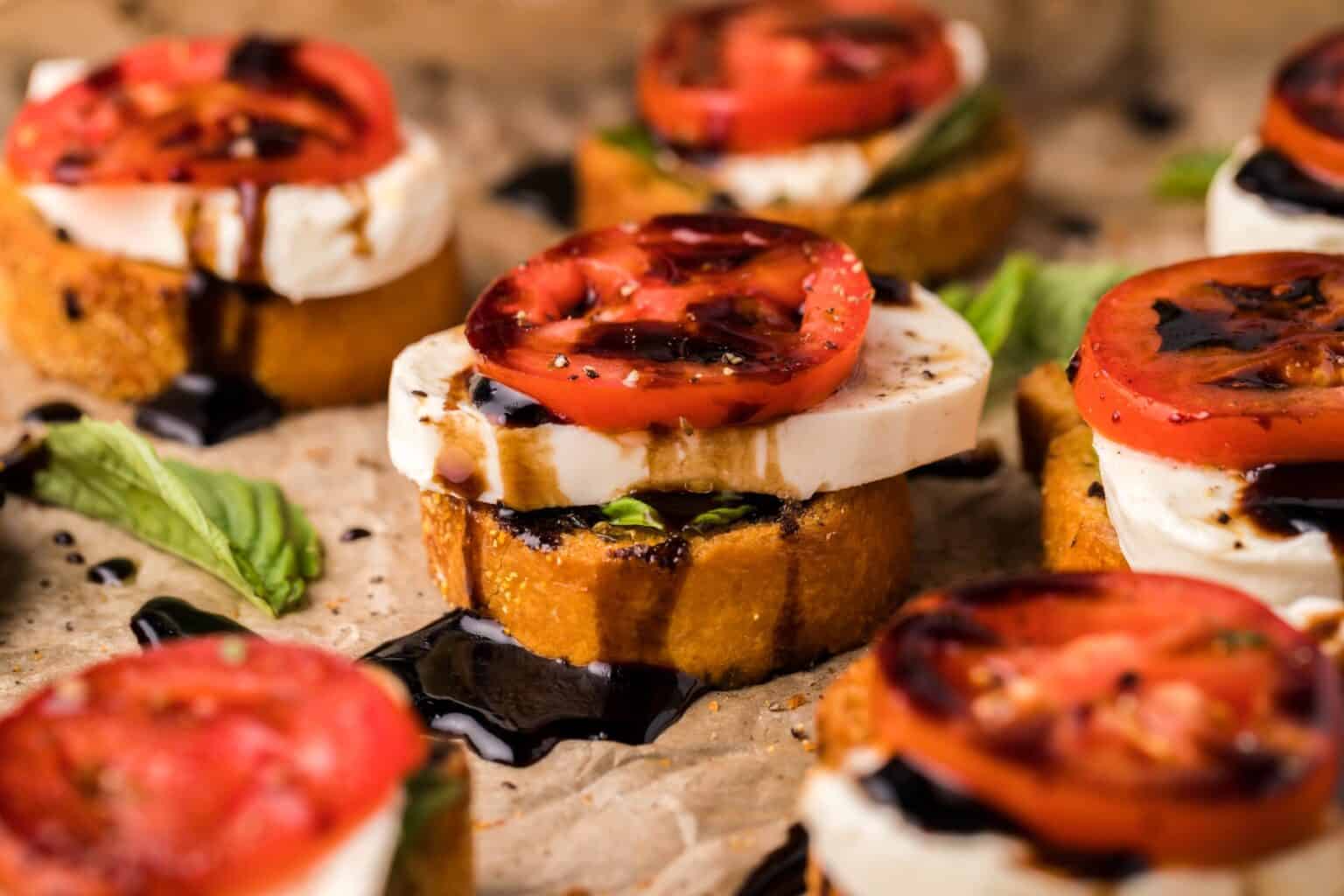 Close-up of caprese bruschetta on parchment paper. Each piece features a toasted baguette slice topped with fresh basil, mozzarella, a tomato slice, and a drizzle of balsamic glaze.