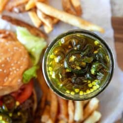 Overhead of candied jalapenos in a jar and in the background in a burger and fries.