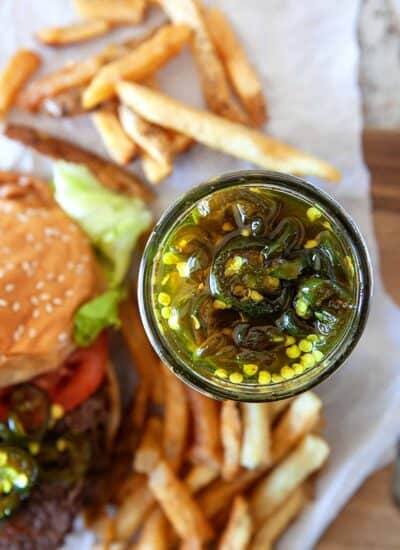 Overhead of candied jalapenos in a jar and in the background in a burger and fries.