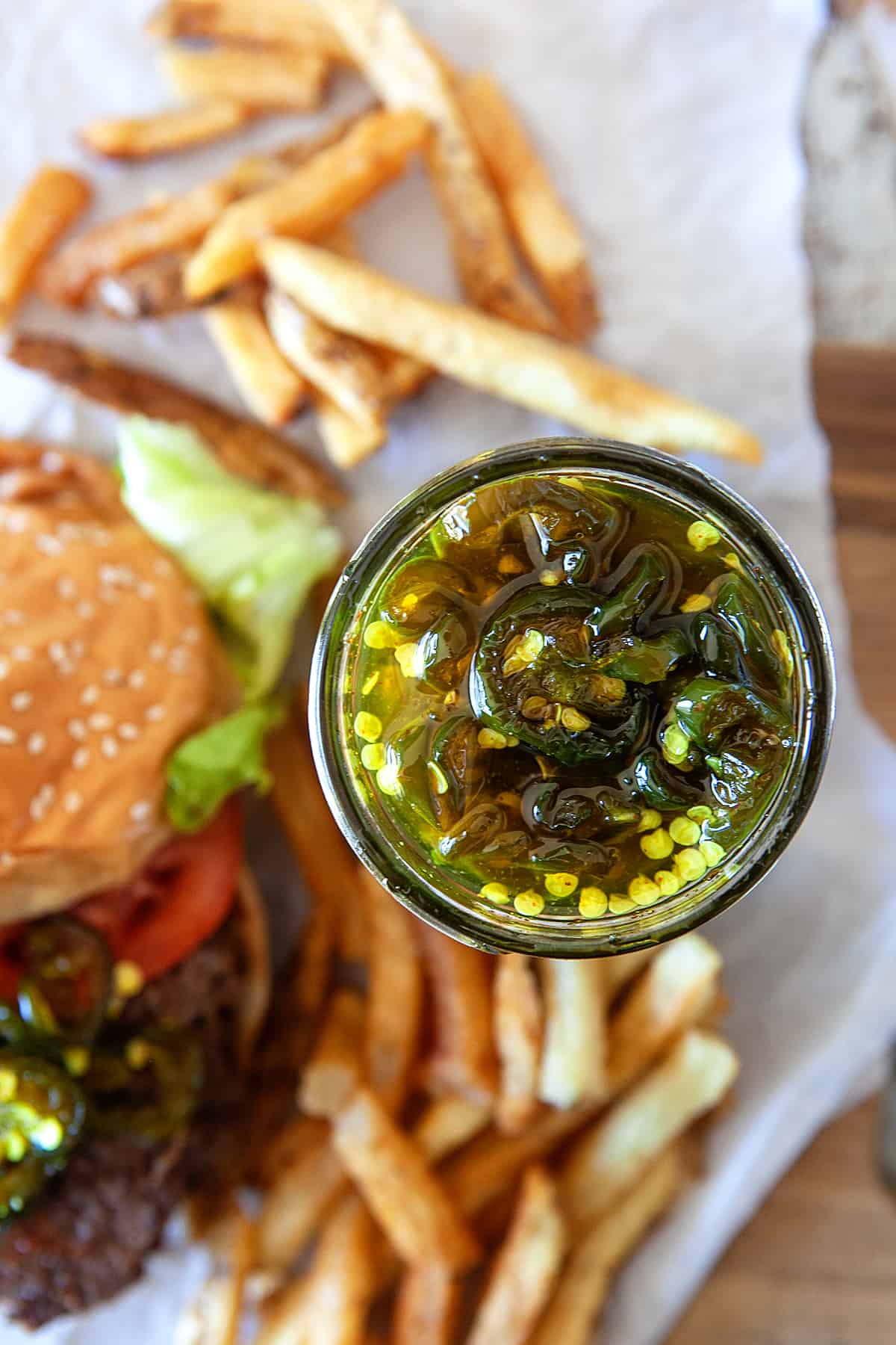 Overhead of candied jalapenos in a jar and in the background in a burger and fries. 
