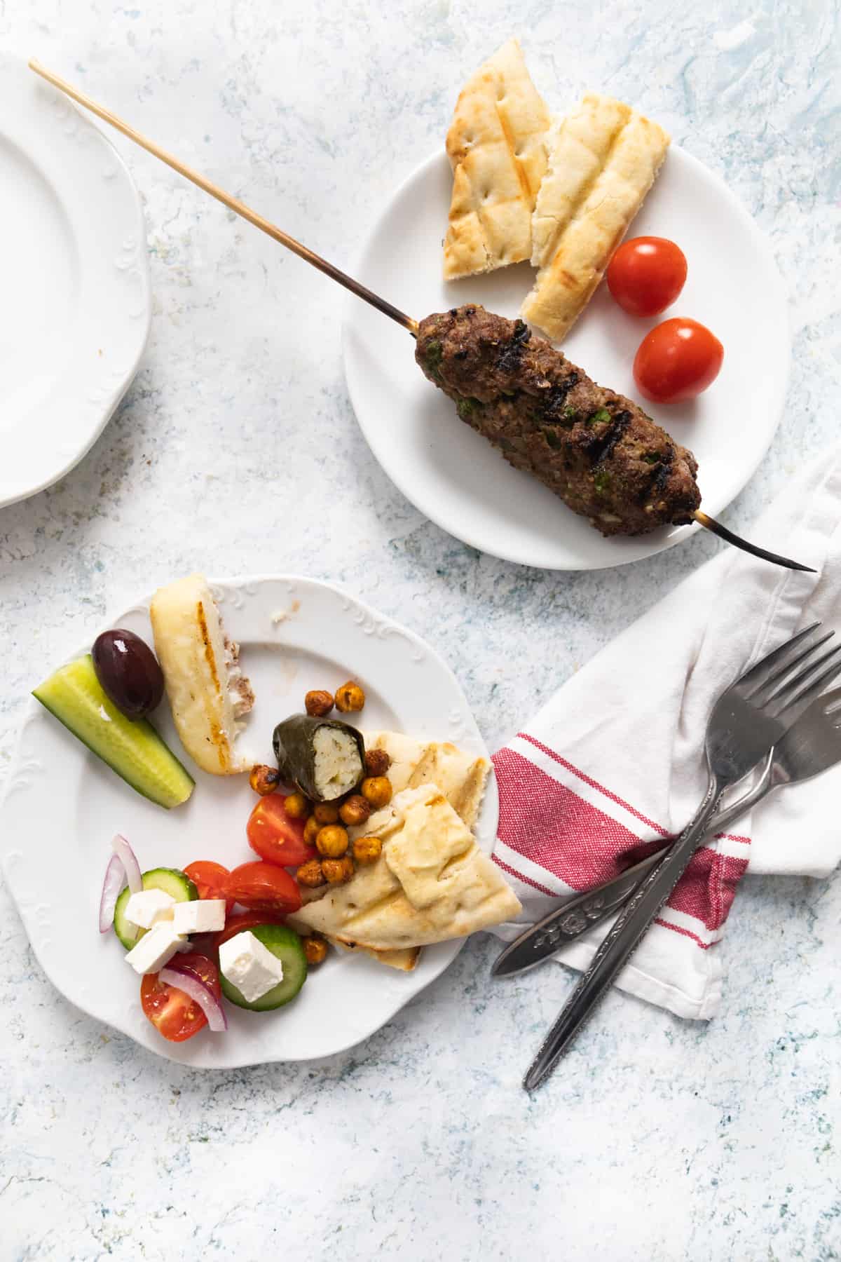 Top view of 2 small plates with one kofta, cherry tomatoes, cucumber, hummus.