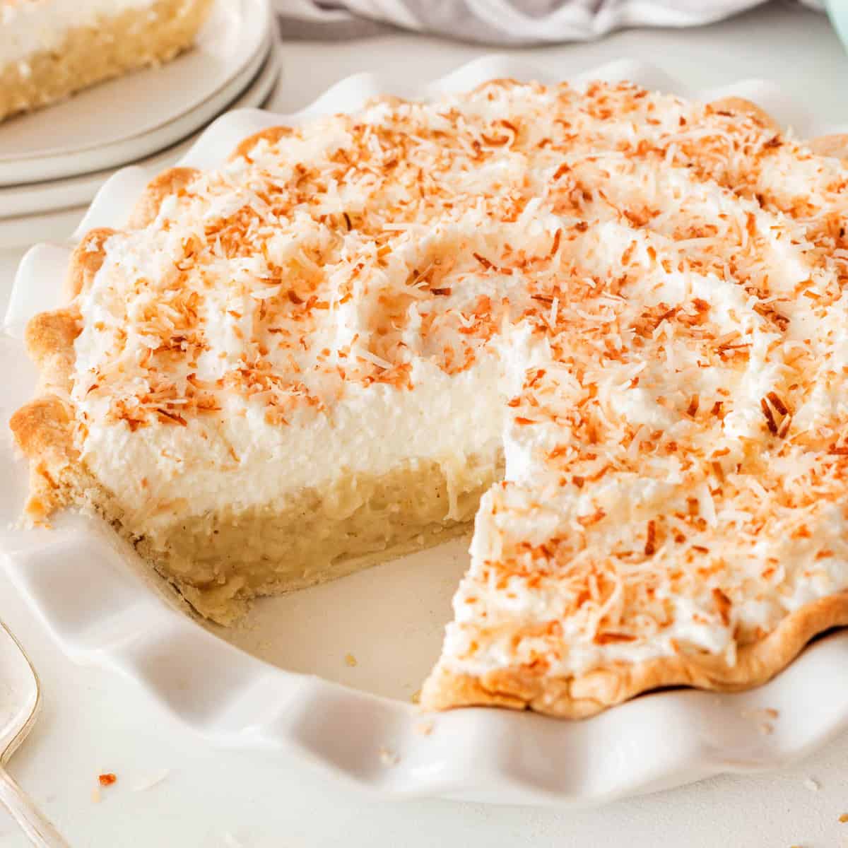A square photo of coconut cream pie with a slice missing. 