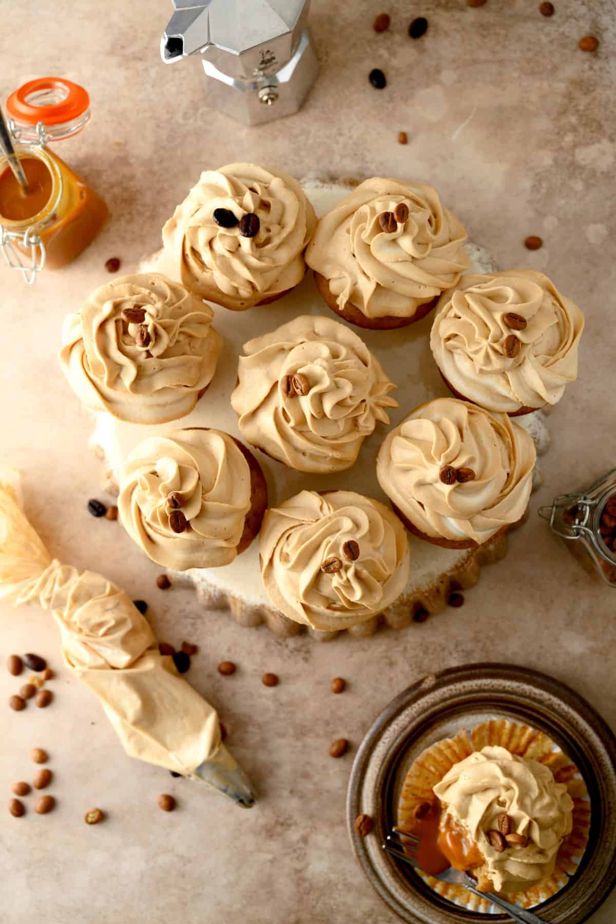 Overhead shot of iced coffee cupcakes with coffee beans on top with coffee beans, caramel in a jar and and an icing bag around them. 