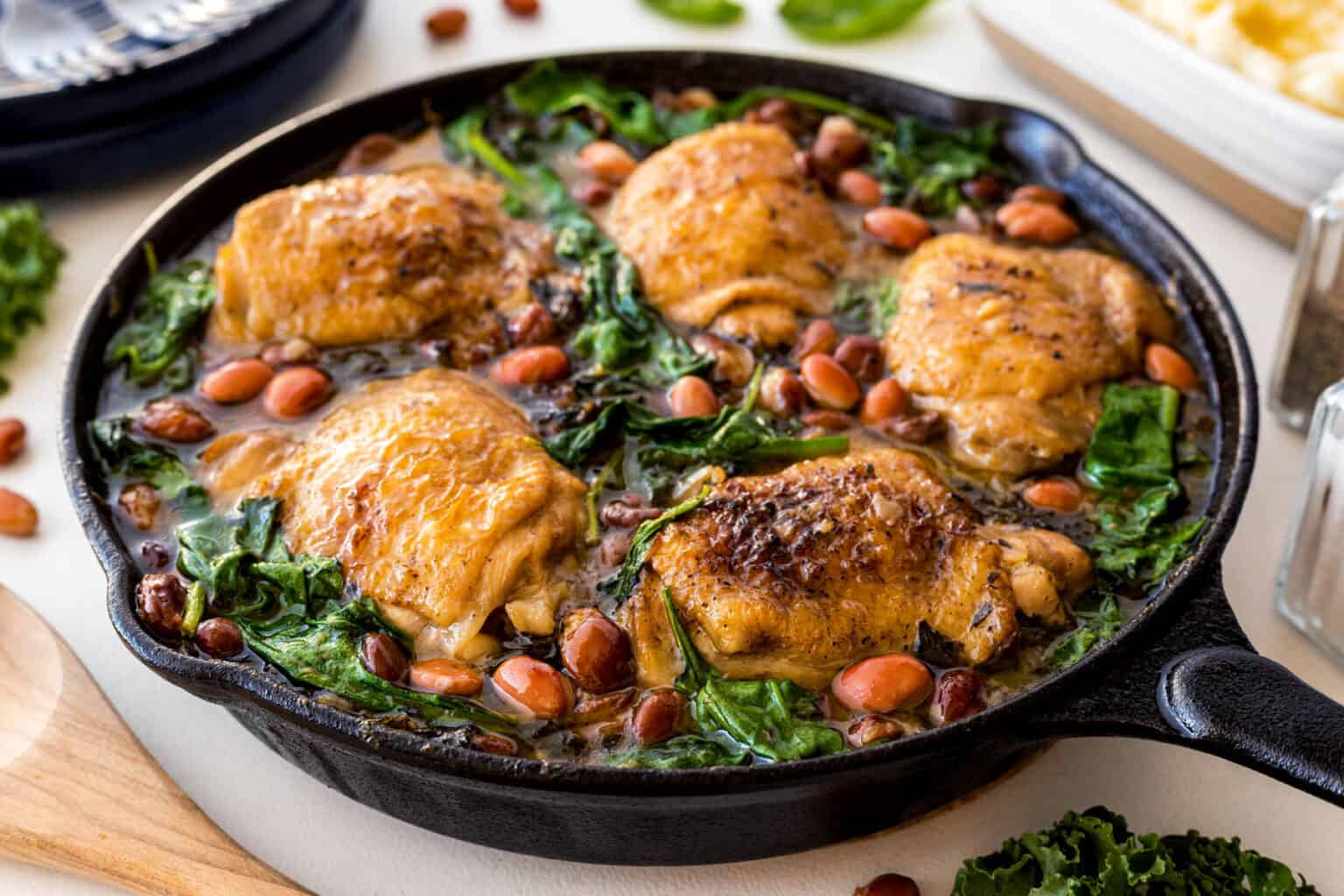A cast-iron skillet filled with baked chicken thighs, surrounded by cooked kale and pinto beans in a savory sauce. The dish is placed on a light-colored surface, and various kitchen items can be seen in the blurred background, including a wooden spoon and a dish.