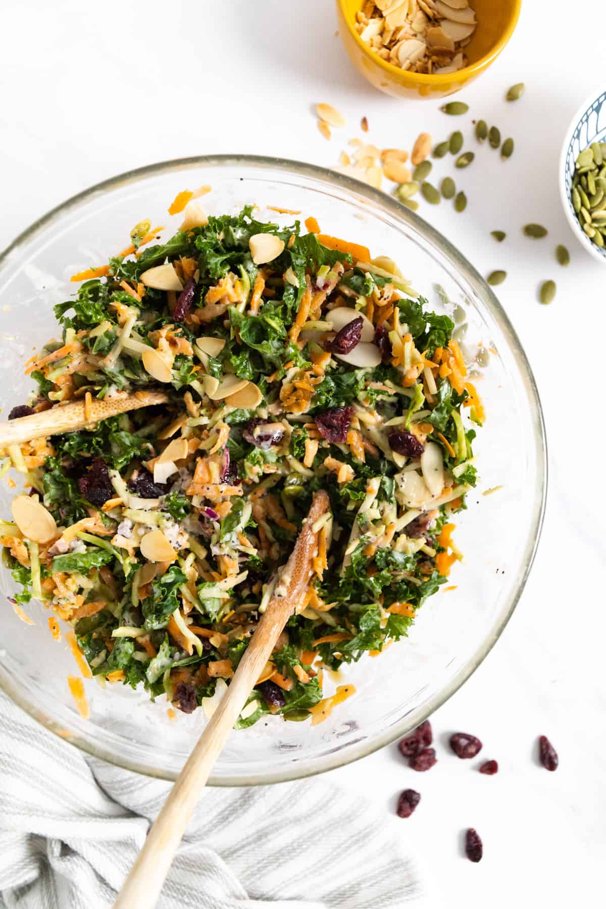 Kale salad in a transparent bowl with wooden spoon tucked in and some almond and pumpkin seeds on the side.