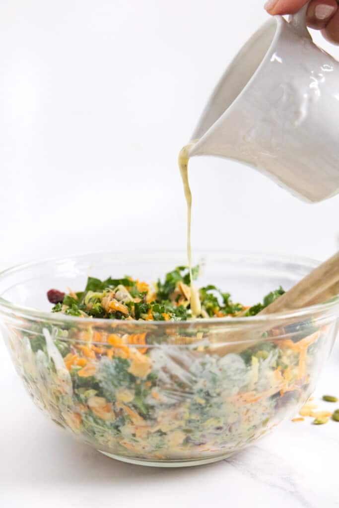 small white pitcher pouring a creamy dressing over a salad in a transparent mixing bowl.