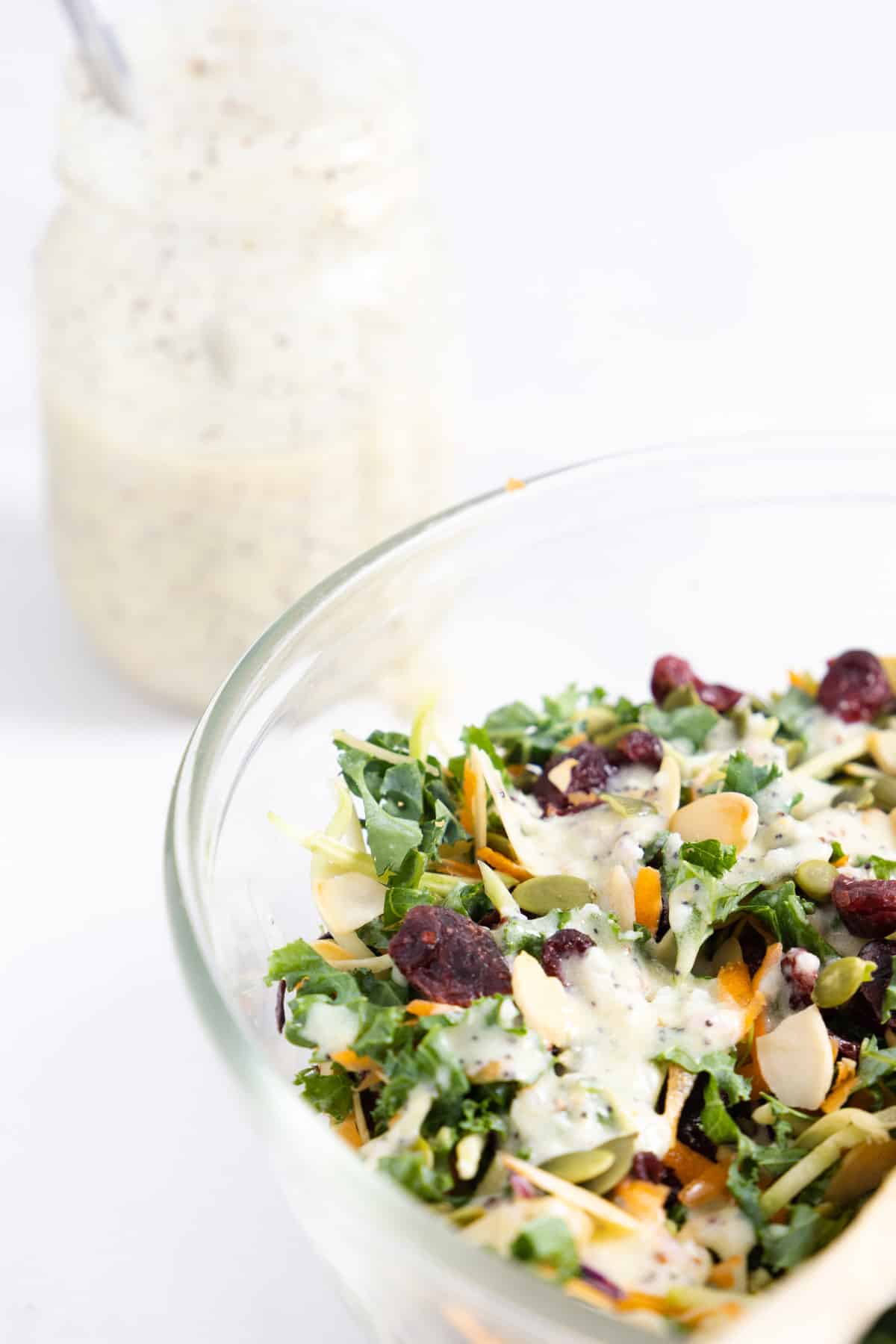 Partial view of a transparent bowl with kale salad in it and in the background a jar with a white creamy dressing. 