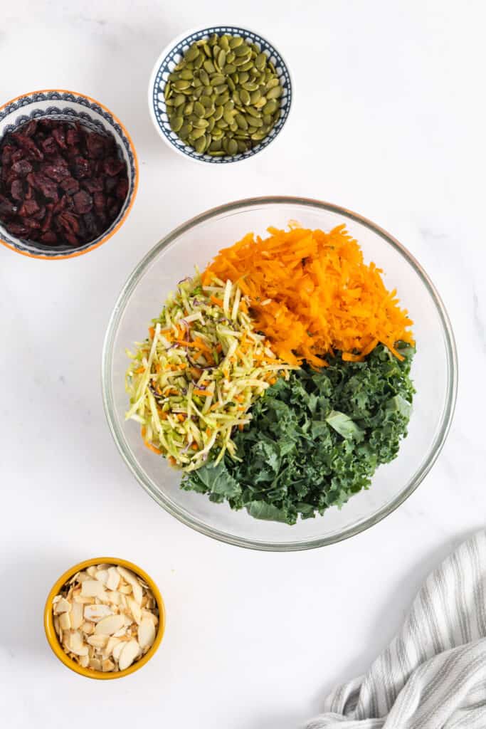 shredded carrots, kale and broccoli slaw in a glass bowl and 3 ramekins with each sliced almonds, dried cranberries and pumpkin seeds. 