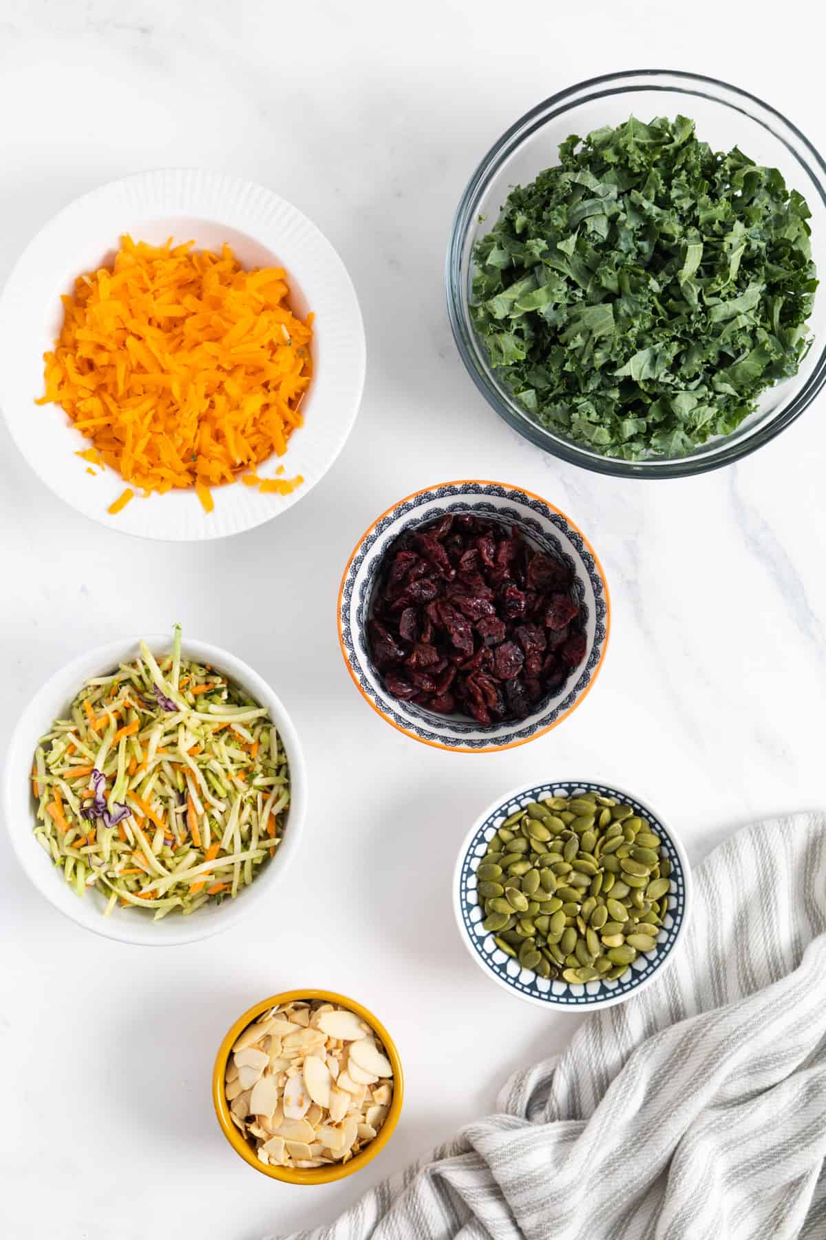 Ingredients on a marble slate: all in a small bowls: kale, slaw, carrots, dried cranberries, pumpkin seeds, almond. 