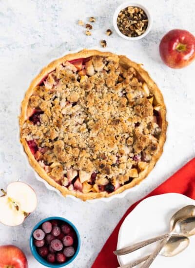 top view of a baked apple crumble pie with ramekins on the side with walnuts and cranberries. A halved apple on the side.
