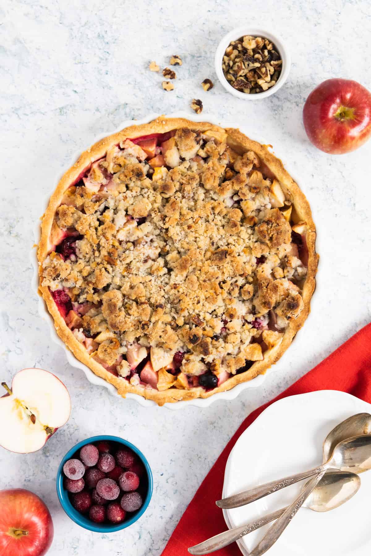 Top view of a baked apple crumble pie with ramekins on the side with walnuts and cranberries. A halved apple on the side. 