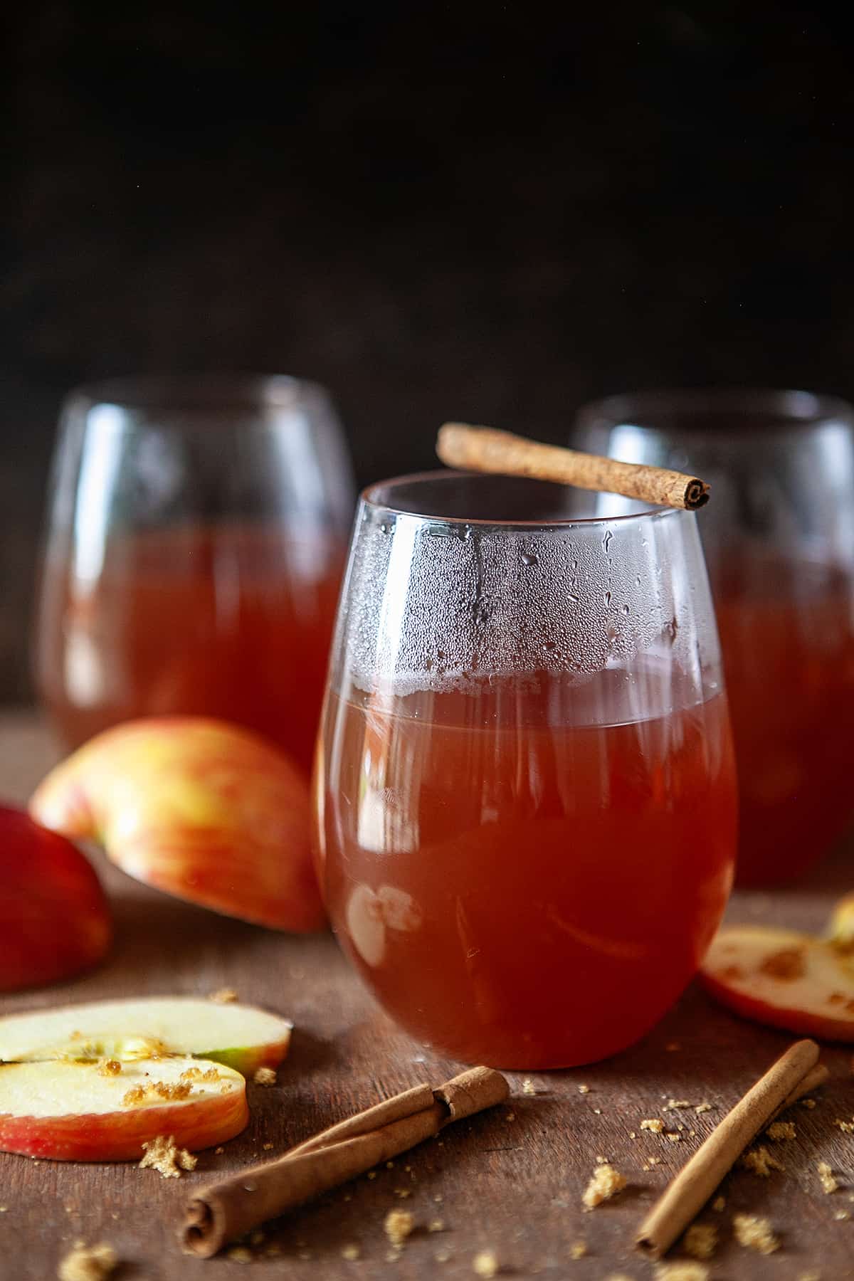 Front view of apple cider in glasses with apples around, the front one has a cinnamon stick on top. 