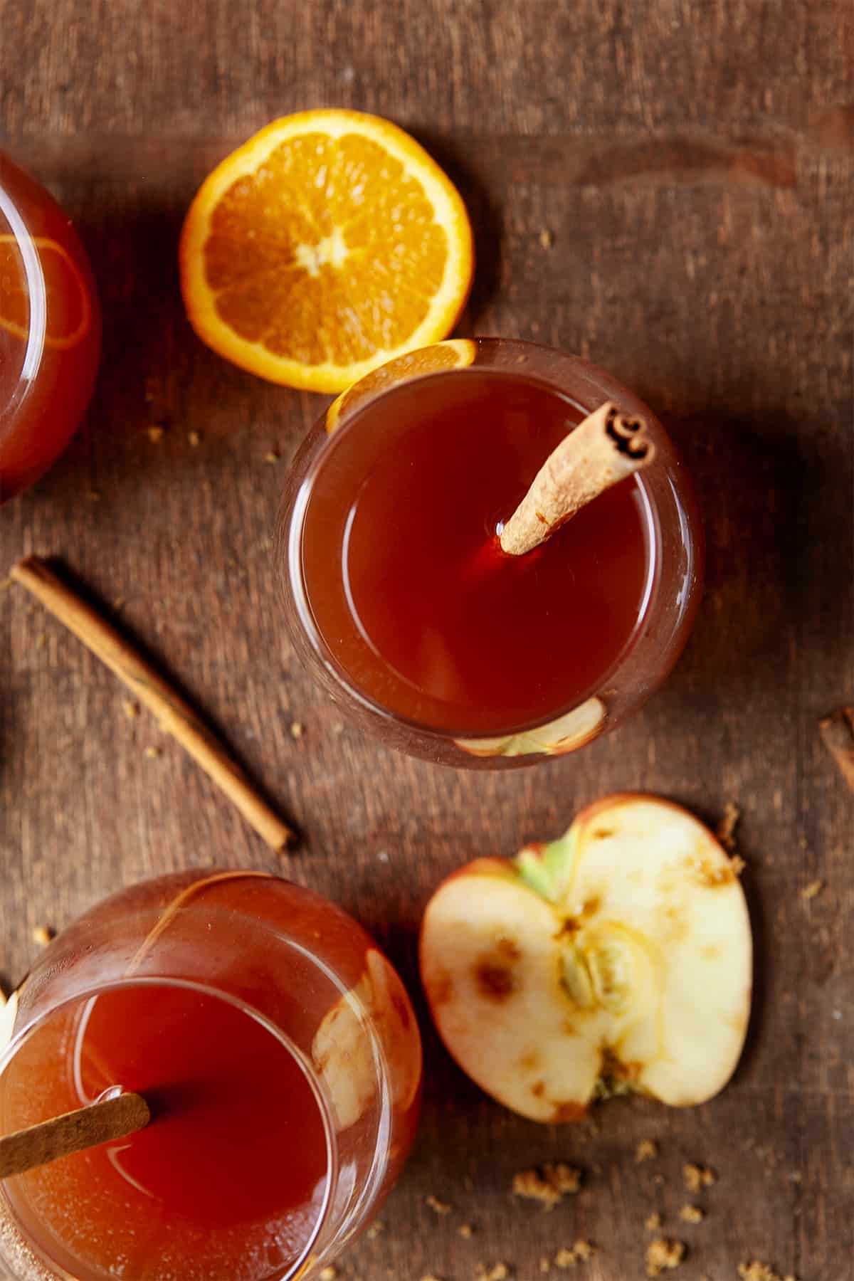 Overhead of Apple Cider in glasses with a slice of apple on the wood below and a cinnamon stick in the glasses and on the wood. 