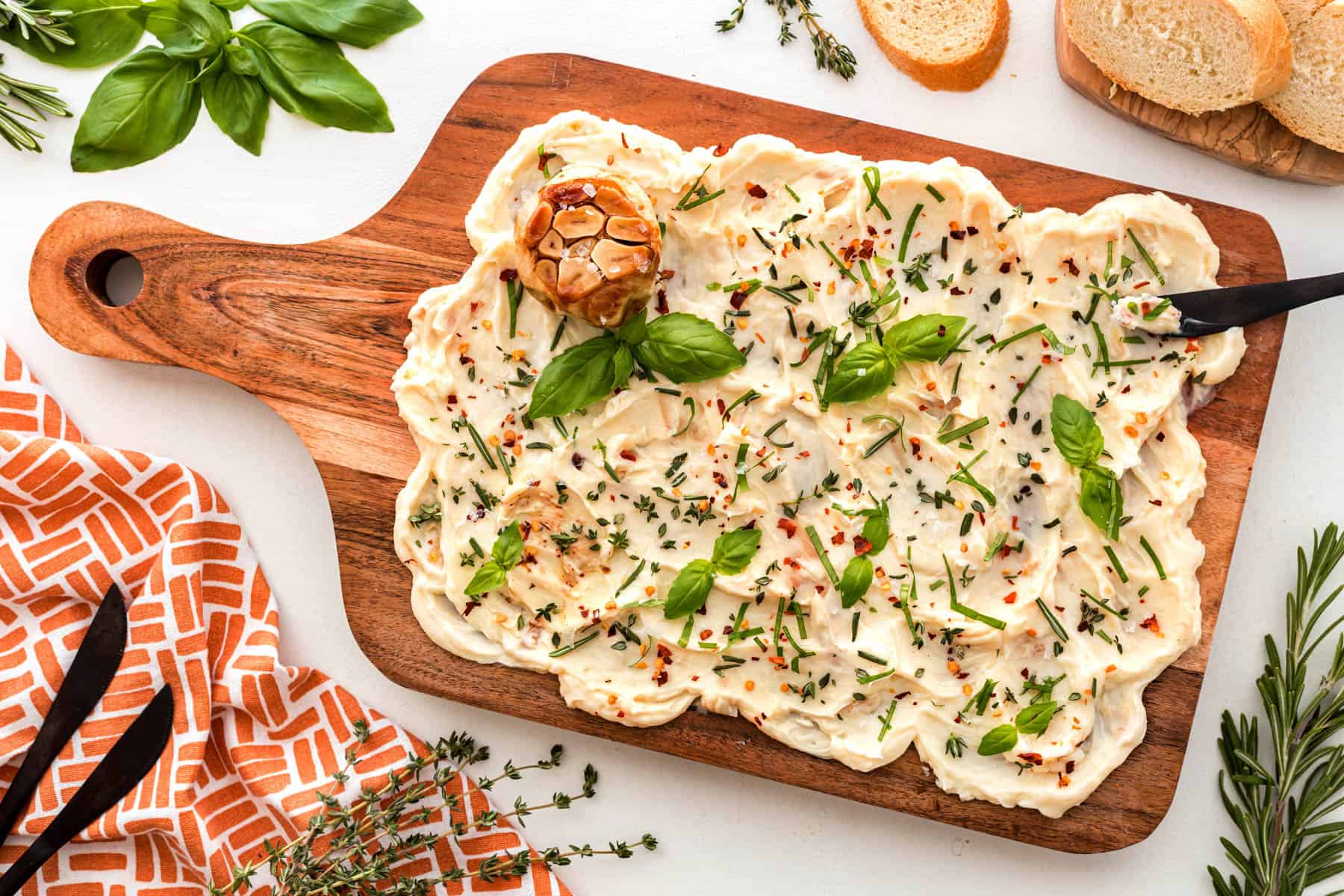 Horizontal overhead of a butter board with fresh herbs and red pepper flakes. Sliced bread and basil at the top of the picture. 