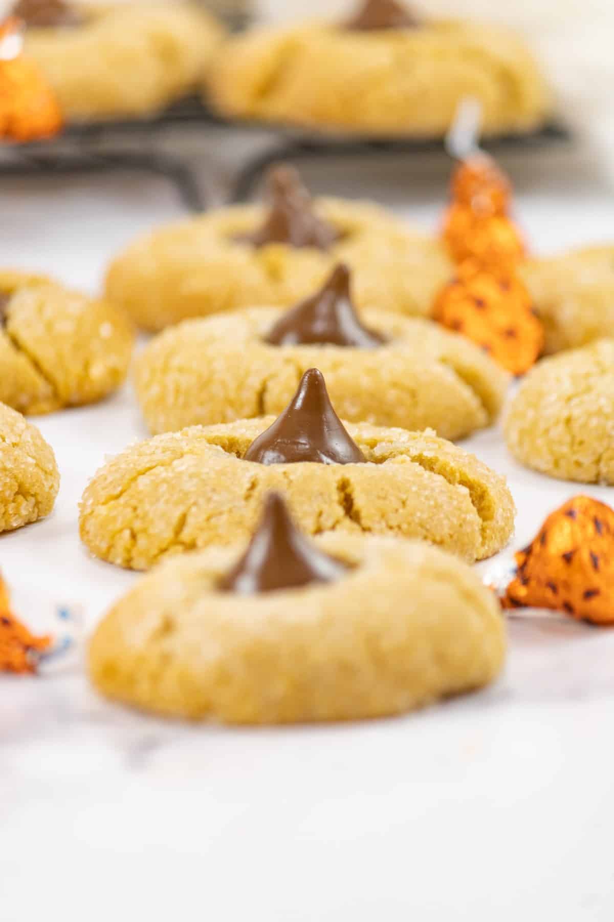 Peanut Butter Blossom Cookies in a row with orange wrapped kisses scattered around. 