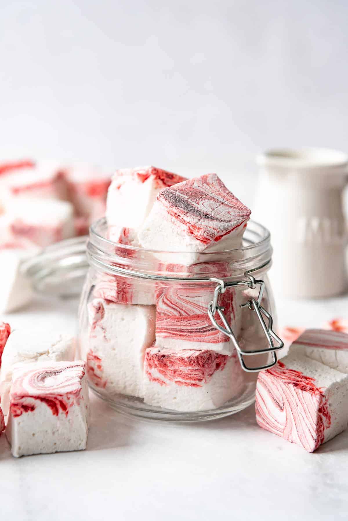 Homemade Peppermint Swirl Marshmallow squares in a small glass jar and also around the jar on the tabletop. 