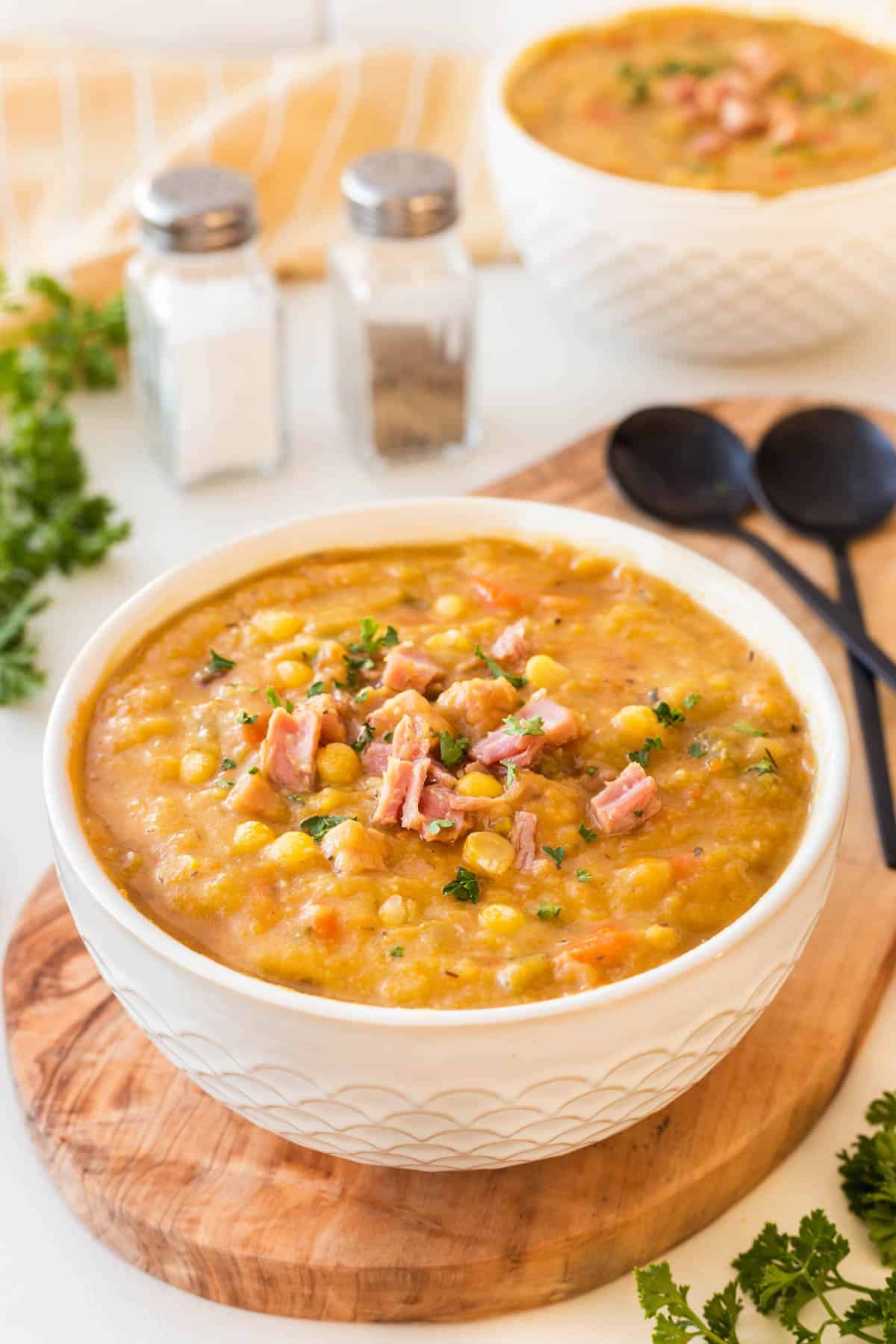 Split Pea Soup with Ham in a white bowl on a board with black spoon, salt and pepper in the background. 