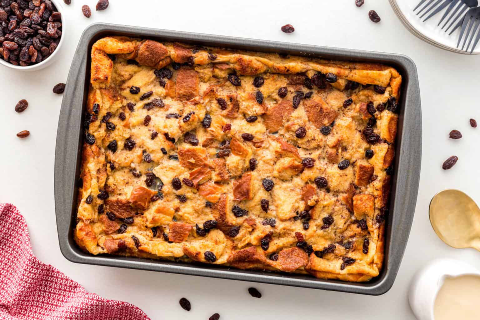 A freshly baked bread pudding in a rectangular pan, topped with raisins. Beside the pan are scattered raisins, a red cloth, a white dish, and a gold spoon.