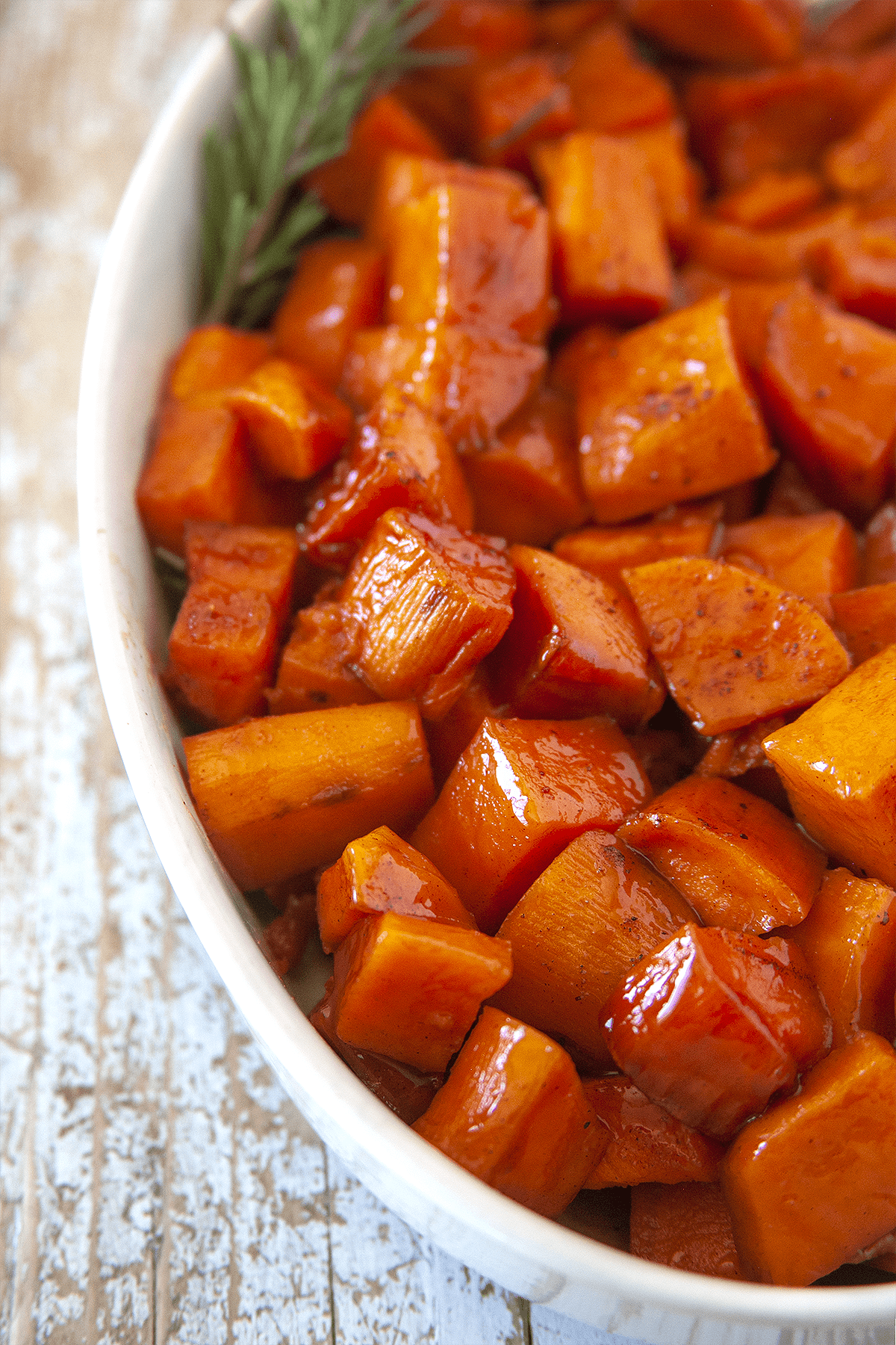 Close-up shot of candied yams in a white oval casserole with a sprig of rosemary. 