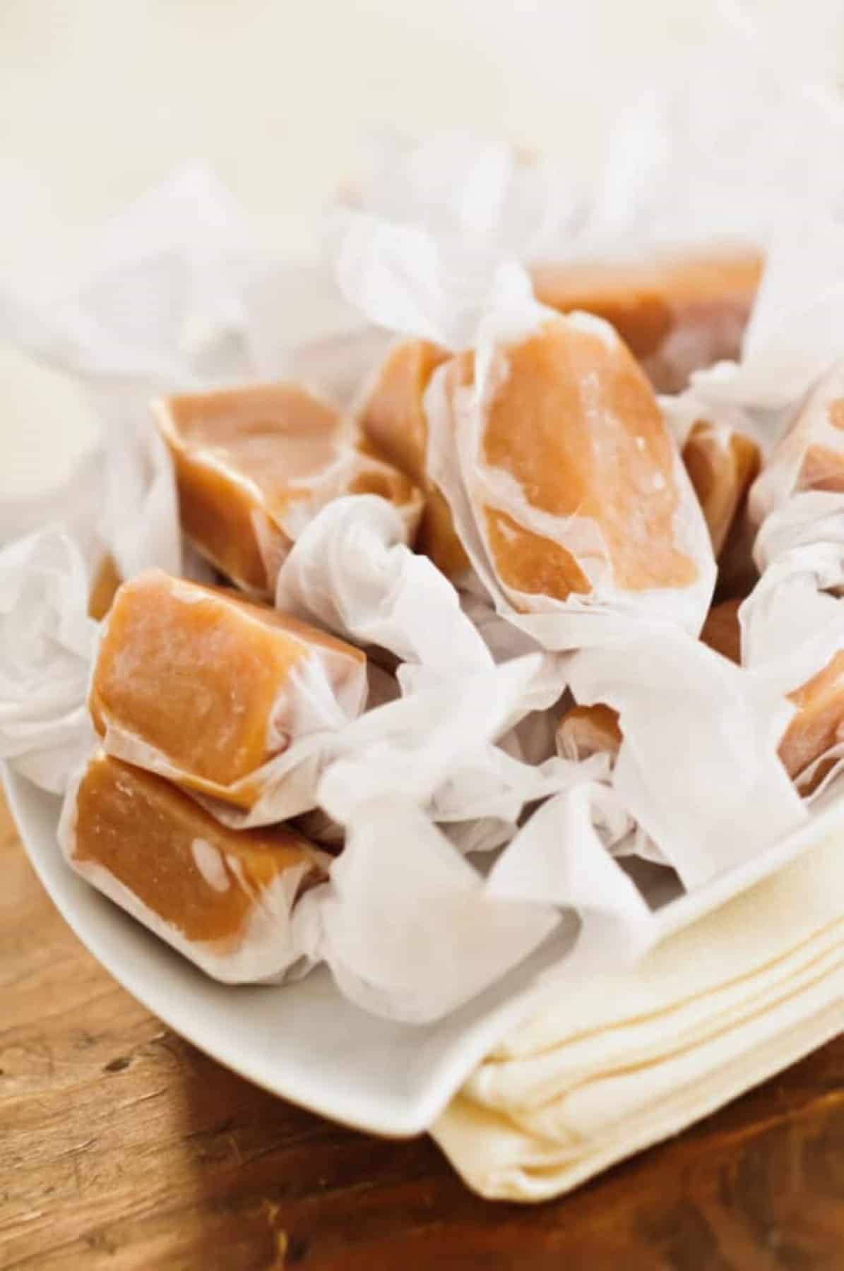 Homemade Christmas Caramel wrapped in wax paper in a white bowl on a wooden tabletop.