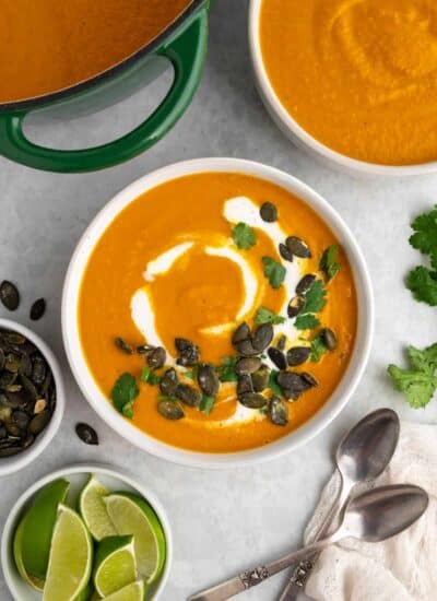 Bowl of pumpkin soup with toppings and also the pot of soup, another bowl. Pepitas and lime wedges in small bowls.