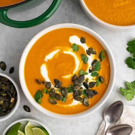 Bowl of pumpkin soup with toppings and also the pot of soup, another bowl. Pepitas and lime wedges in small bowls.