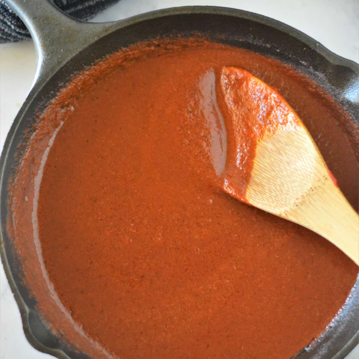 Square photo of enchilada sauce in a cast iron pan with a wooden spoon. 