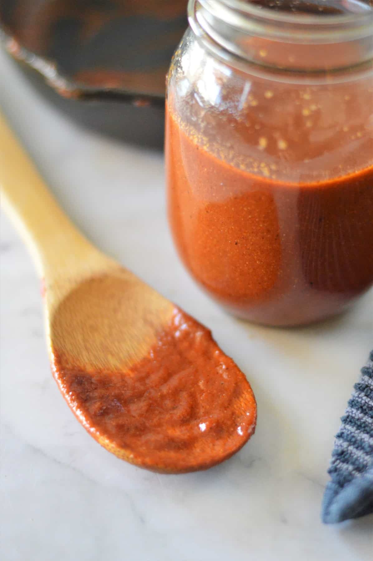 Homemade enchilada sauce in a mason jar with a wooden spoon covered in sauce on the counter beside the jar.