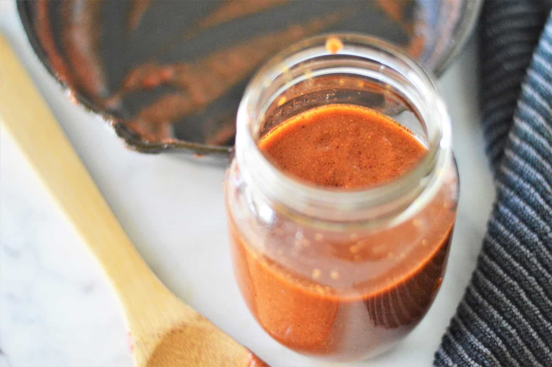 Easy enchilada sauce in a mason jar on a white counter with a wooden spoon beside the jar.