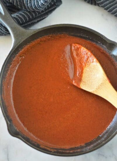 Square photo of enchilada sauce in a cast iron pan with a wooden spoon.