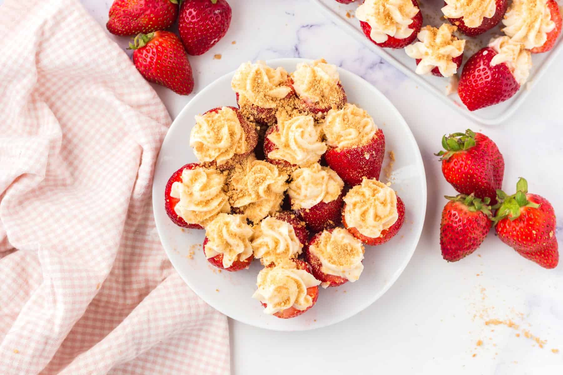 Horizontal photo of cheesecake stuffed strawberries on a round white plaste with fresh strawberries around and a checkered pink tea towel. 