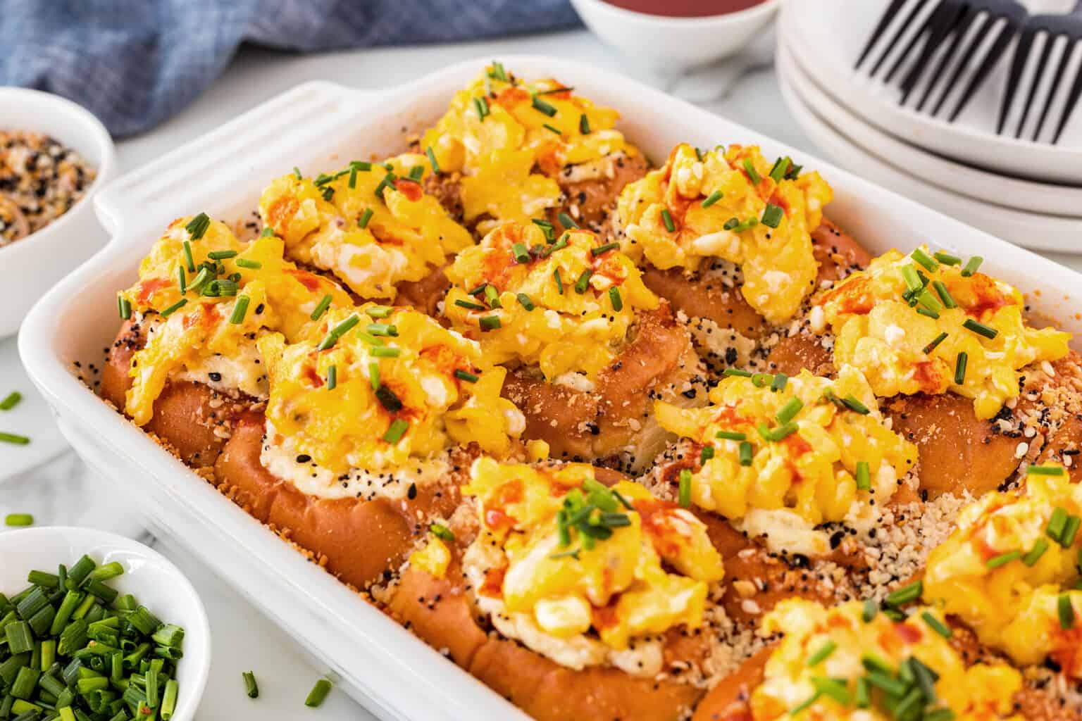 A casserole dish filled with golden, cheesy rolls topped with chives and a sprinkle of seasoning, accompanied by fresh chives and plates.