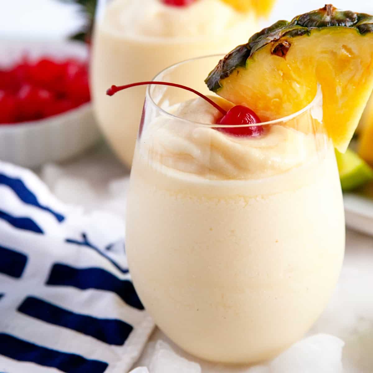 Square photo of a pina colada in a round glass garnished with pineapple and a cherry. 