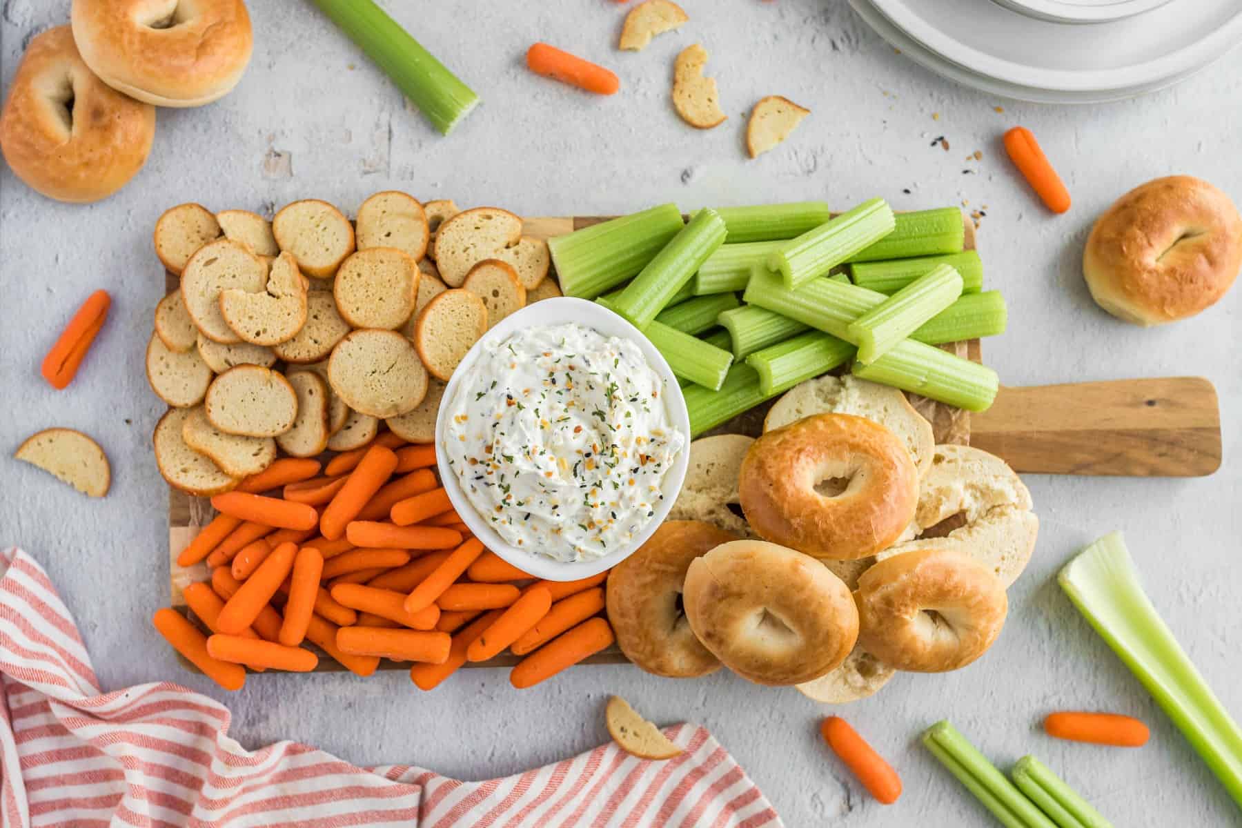 Horizontal picture of a wooden platter of carrots, bagels, crostini, celery and the dip in the middle. 