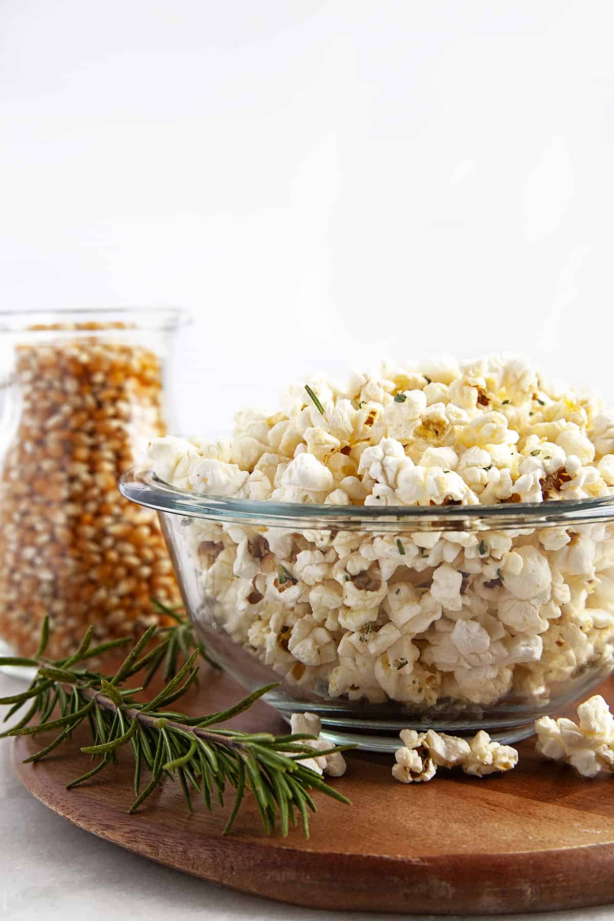 Front view of popcorn in a glass bowl with kernels in the background in a jar and a sprig of rosemary up front. 