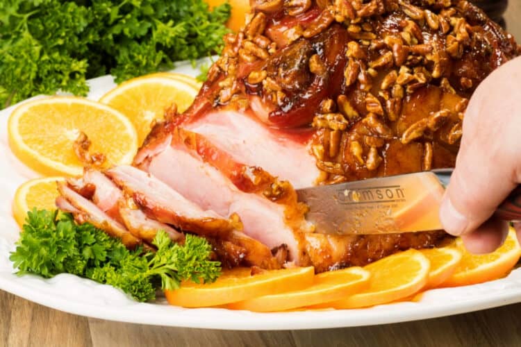 Ham being sliced on a plater with oranges and parsley around it.