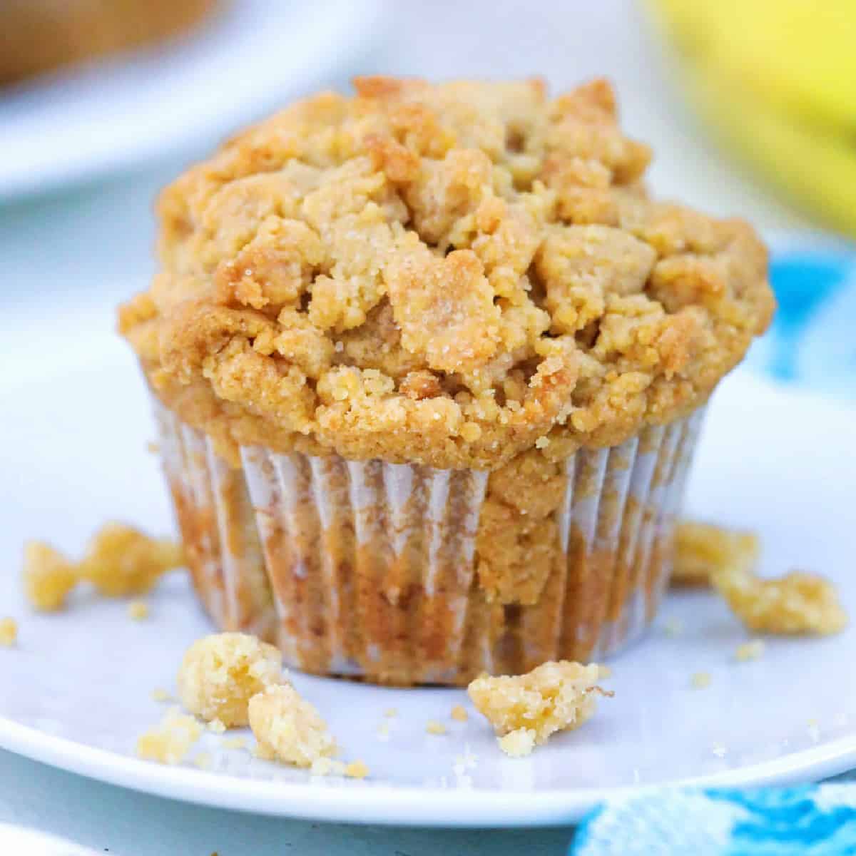 Square image of a muffin on a plate with crumble topping around the muffin. 