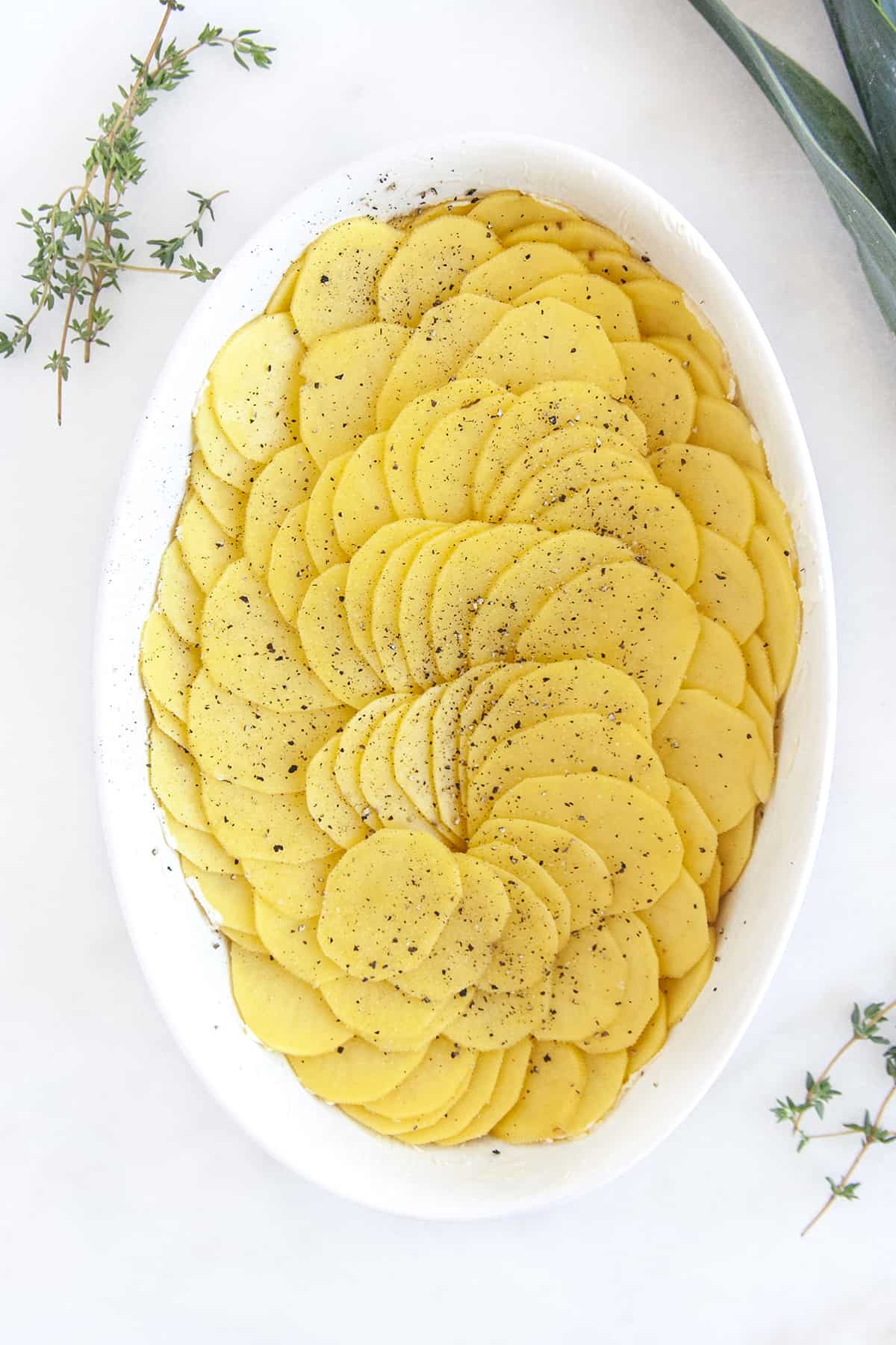 Slices of potatoes laid out in a circular fashion in an oval baking dish with salt and pepper on top. 