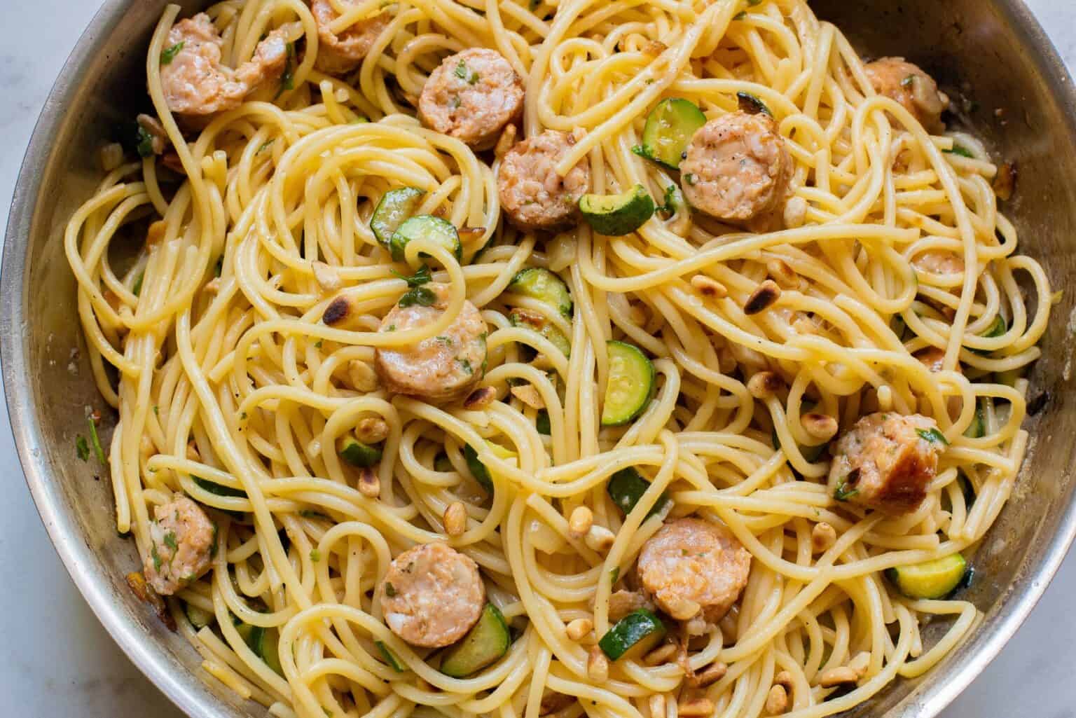 A close-up of a pan containing spaghetti mixed with slices of sausage, zucchini pieces, and pine nuts. The dish appears well-cooked and garnished, with the ingredients evenly distributed throughout the spaghetti.